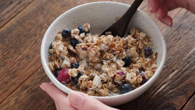 Eating granola with almond milk. Female hands mixing oat coconut granola with berries and almond milk on a rustic wooden table background. Healthy breakfast food