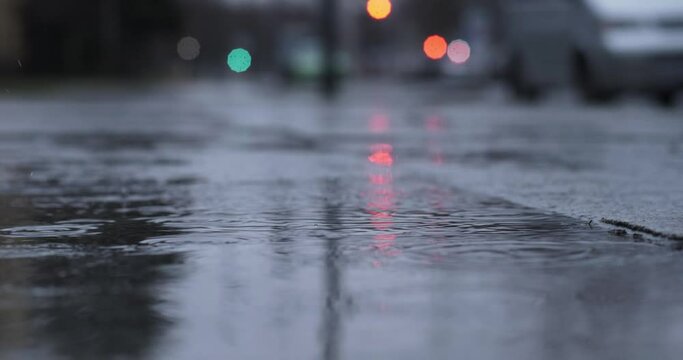 Raindrops hitting a wet street during cold rainy weather. Lights reflecting in the water. Dark moody evening look and no people in the city.