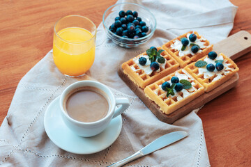 Waffles with cream and blueberries, a cup of coffee and orange juice on the table on a cloth napkin. Top view