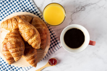 Several small croissants on blue checkered cloth along with a cup of coffee, orange juice and rich marmalade and butter spread. 