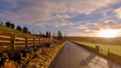 A moody image taken along a rural road with a wooden fence and drystone wall leading into the distance. All lit by a low sun with soft clouds and blue sky.