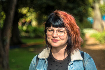 Portrait young caucasian woman with black and red hairstyle, jean jacket and vintage glasses looking at camera smiling and laughing naturally and spontaneously. Green trees and park in the background