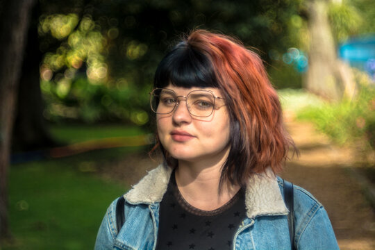 Portrait of a young caucasian woman with black and red hairstyle, jean jacket and vintage glasses looking at camera with serious expression. Green trees and park in the background