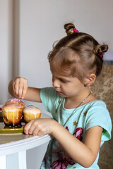 Fototapeta premium Little girl decorates Easter cakes. At home in the kitchen, the baby helps to prepare for Easter. A girl sprinkles cakes with colorful dragees.Keen child