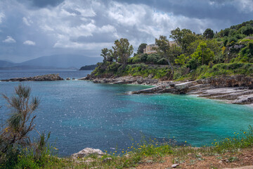 Beautiful landscape - sea lagoon with turquoise calm water, stones and rocks on the beach, clouds on the sky, green trees and bushes, mountains on the horizon. Corfu Island, Greece, Kassiopi village