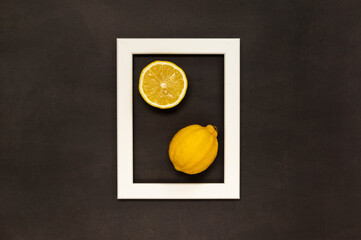 Whole and half of lemon put in white photo frame on black background. Fruit flat lay concept. Color minimalism of yellow, white and black.