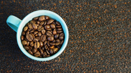 a blue mug filled with whole roasted coffee beans stands on a black textured background top view