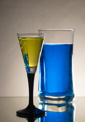 Highball glass filled with aqua and blue dye ink, Liqueur glass filled with aqua and yellow dye. Isolated objects in the studio with refelcting surface and gradient background 