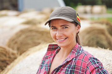 Young woman in shirt and cap smiling, hay stack rolls behind her