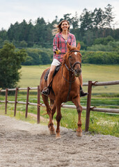 Young woman wearing shirt riding brown horse in sand paddock by wooden fence, hair moving in air because of speed