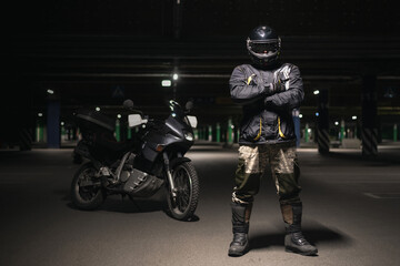 A motorbiker is standing with crossed hands on a dark parking.