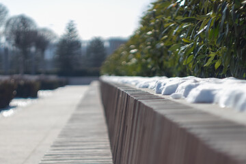 blurred long park bench in spring with snow and green shrubs on a bright sunny day