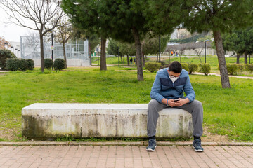 Fototapeta premium Young boy wearing face mask due to covid19 using his cellphone while sitting in a bench in the park.