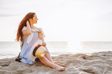 Naklejka premium Young woman in dress and straw hat sit alone on empty sand beach at summer sunset on sea shore and and smile. Space for message.