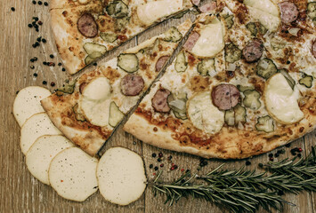 Super cheesy pizza with hunting sausages, pickles and melted cheese. sprigs of rosemary lie nearby