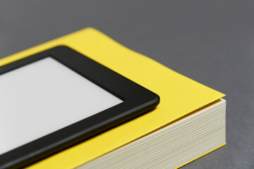 Electronic reader with blank screen and closed yellow book on gray background