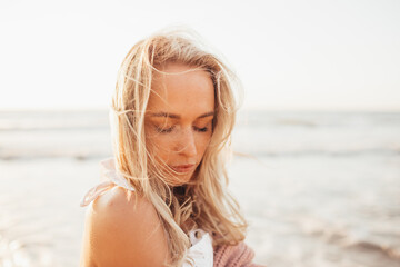Headshot of beautiful blonde slim girlwearing white dress and cardigan on beach of sea or ocean against the sun. Youth, vacation, travel, fashion.