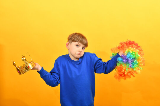 The boy holds a clown wig in one hand and a golden crown in the other. The choice between control and play.