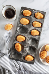 Top down view of freshly baked madeleine cakes with a steaming cup of coffee on white marble background