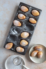 Flat lay of a tin of freshly baked madeleine cakes, some on a plate and a sieve with icing sugar