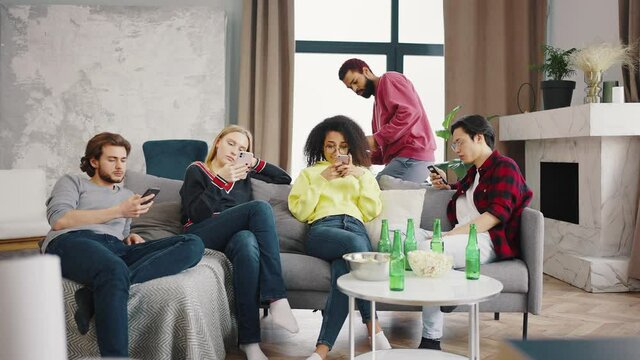 Group of young multiracial diverse friends using smartphone with mutual disinterest towards each other in loft appartment. Phone addicted young multiethnic people phubbing each other indoors