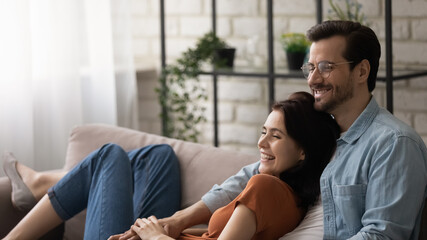 Warm relationship. Laughing young wife rest on comfy couch at home snuggle up to loving husband. Couple in love relaxing at living room having fun dreaming looking at distance enjoy nice conversation