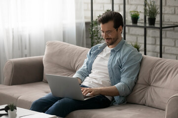Inspired with job. Smiling young man wearing glasses sit on cozy couch hold laptop on knees type email to client surf internet check news. Happy guy freelancer work online from home at convenient time