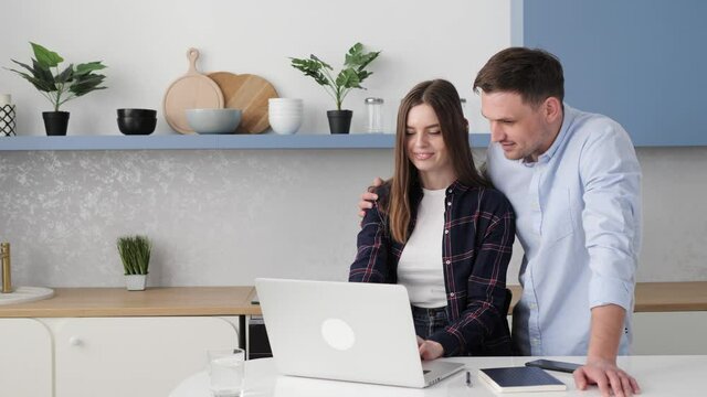 Young caucasian couple of woman and man sitting at home kitchen looking to computer laptop screen smiling laughing pointing to screen excited cheerful happy. Couple buying products or services online.