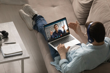 Young man trainee sit on couch in wireless headphones listen to indian woman trainer on pc screen. Focused guy student meet with biracial female teacher on remote consultation. Over shoulder top view - Powered by Adobe