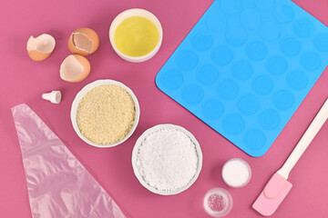 Ingredients for making homemade French Macaron sweets including powdered sugar, ground almonds, egg white, salt and baking tools like mold mat and icing pipe on pink background