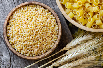 Raw couscous in bowl on wooden background with wheats, uncooked food concept