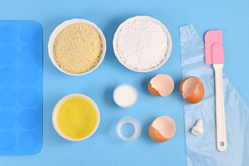 Ingredients for making homemade French Macarons sweets including powdered sugar, ground almonds, egg white, salt and baking tools like mold mat and icing pipe on blue background