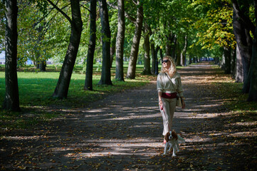 young blonde woman in light casual clothes walks with a Cavalier King Charles Spaniel dog in the historical center of the small town of Kronstadt on a sunny fine day along the cobblestones