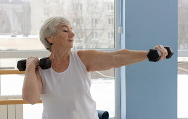 An older athletic woman does a dumbbell exercise in a light gym. Active old age, sports for the...