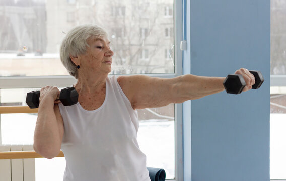 An Older Athletic Woman Does A Dumbbell Exercise In A Light Gym. Active Old Age, Sports For The Elderly Concept