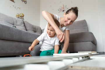 Cute adorable caucasian toddler boy kid sit on floor and help mom assembling furniture shelf with power screwdriver tool. Young adult mum with funny little child enjoy playing at home indoors