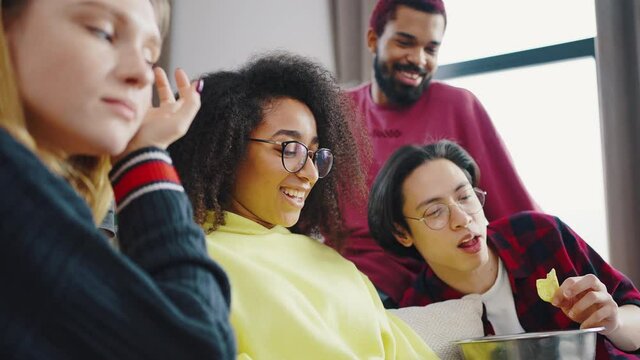 Interacial group of friends has fun sitting on the couch with an indoor laptop. Boys and girls eat chips and watch movies indoor. People and gadgets concept