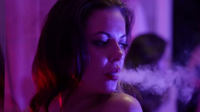 sensual woman is smoking to window at night, inhaling and exhaling smoke of e-cigarette
