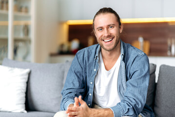 Portrait of an attractive charismatic caucasian blue-eyed guy in a denim shirt sitting on a sofa in...