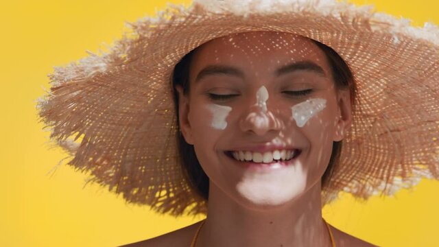 Close-up beauty portrait of young attractive dark-haired caucasian woman in a straw hat applies sunscreen on her nose laughing for the camera | Sunscreen application shot for face care commercial