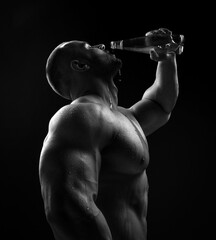 Brutal sweaty strong young man athlete with naked upper body standing drinking water from bottle...