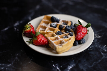 Waffles with strawberries and blueberries presented covered in powered sugar on a granite counter top