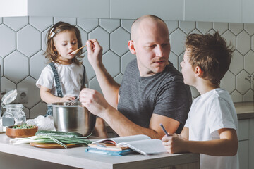 Father whips omelette with whisk, helps his son do his homework and feeds his daughter in kitchen. Man doing chores.