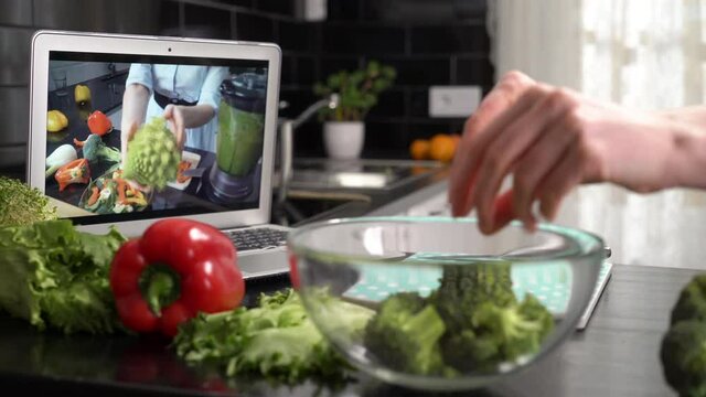 A young woman watching a online stream broadcast video blog tutorial about tasty and healthy food using laptop and cooking a meal at the home kitchen