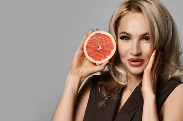 Portrait of smiling surprised young pretty blonde woman holding half of fresh ripe grapefruit in hand looking at camera over grey background. Healthy eating and balanced diet concept