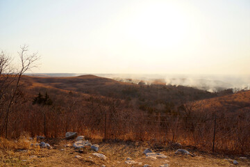 Fototapeta premium Wildfires in the distance burn brush and dry grass in the hills and fills the valley with smoke. A barbed wire fence in the foreground separates pastures on a ranch.