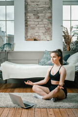 The young woman goes in for sports at home. Cheerful sportswoman with blond hair meditates in lotus...