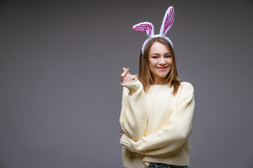 smiling young caucasian girl, blonde with bunny ears, shows a mini heart with fingers and looking at camera isolated over gray background