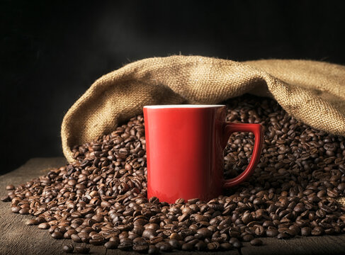 Red mug with hot coffee on the background of a heap of coffee beans spilled from a bag on the table