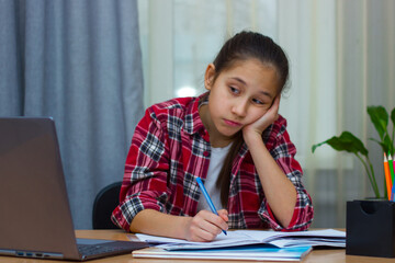 Sad brunette girl dressed in a red shirt, sitting at home studying, writes homework, communicates on the Internet. 